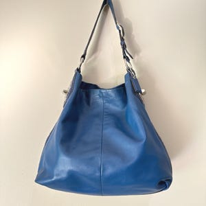 May include: A deep blue leather hobo bag featuring a single adjustable shoulder strap with silver-toned metal hardware, including circular rings and a buckle. The bag has a slouchy, unstructured silhouette with a smooth finish and a top zipper closure.