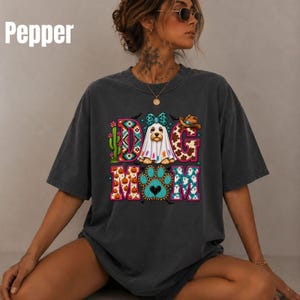 Puede incluir: Una camiseta gris carbón con un estampado gráfico que incluye el texto "DOG MOM". El diseño muestra un perro con disfraz de fantasma, un cactus, un sombrero de vaquero y una huella de pata, decorados con patrones coloridos de calabazas y fantasmas.