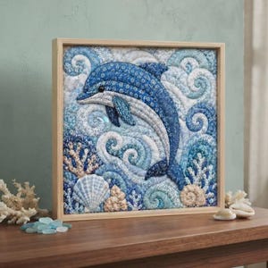 May include: A square framed fabric wall art piece featuring a quilted dolphin in shades of blue and white. The dolphin is surrounded by swirling wave patterns, coral structures, and seashells, all crafted from various patterned blue fabrics.
