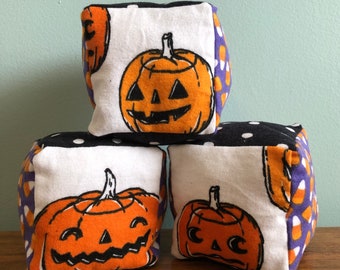 May include: Three fabric cubes with a Halloween theme. Each cube features a different orange pumpkin with a black face and a white background. The cubes are decorated with black and white polka dots, purple and orange candy corn, and a purple background.