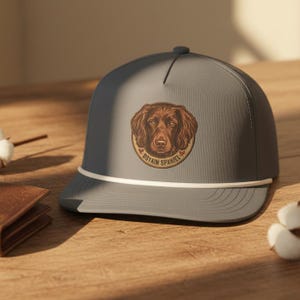 May include: A grey baseball cap featuring a circular patch with a brown Boykin Spaniel illustration and the text "BOYKIN SPANIEL" printed below the dog. The cap includes a white cord detail across the base of the crown and a flat brim.