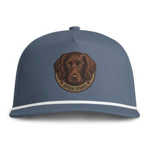 May include: A slate blue baseball cap featuring a white cord trim along the base of the crown. Centered on the front is a circular patch displaying a detailed brown Boykin Spaniel dog illustration with the text BOYKIN SPANIEL printed below.