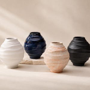 Pode incluir: Quatro vasos esféricos de cerâmica com texturas onduladas horizontais. A coleção inclui um vaso branco fosco, um vaso azul profundo brilhante, um vaso texturizado em tom de pêssego e um vaso preto fosco, todos com aberturas circulares.