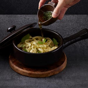 May include: A dark brown ceramic saucepan with a matching lid, containing cooked fettuccine pasta garnished with green herbs. A small matching ceramic pouring vessel is held above, drizzling a golden oil dressing over the pasta dish.