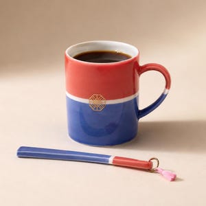 Könnte beinhalten: Ein Keramikbecher mit roter oberer und blauer unterer Hälfte, getrennt durch ein weißes Band mit goldenem geometrischen Emblem. Der Becher ist mit dunkler Flüssigkeit gefüllt. Daneben liegt ein passender blau-roter Keramiklöffel mit rosa Quaste.