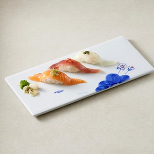 Puede incluir: Un plato de servicio rectangular de cerámica blanca decorado con motivos florales azules, que presenta tres piezas de nigiri sushi cubiertas con salmón, atún y pescado blanco, acompañadas de una pequeña porción de wasabi verde y jengibre.