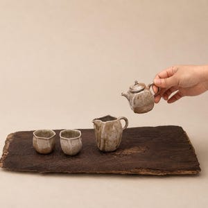 Könnte beinhalten: Ein Keramik-Teeservice bestehend aus zwei kleinen Tassen, einem Milchkännchen oder Krug und einer Teekanne. Die Stücke haben eine gesprenkelte, erdige beige-braune Glasur und eine unregelmäßige, organische Form. Die Teekanne wird in der Luft gehalten.
