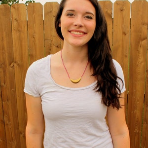 May include: A woman with long brown hair is wearing a white t-shirt with a gold crescent moon necklace. She is standing in front of a wooden fence.