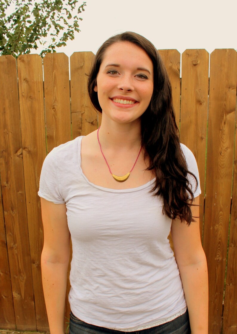 May include: A woman with long brown hair is wearing a white t-shirt with a gold crescent moon necklace. She is standing in front of a wooden fence.