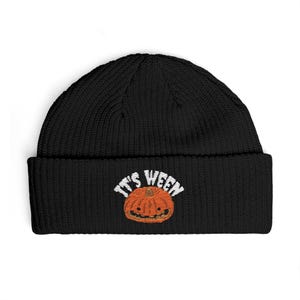 May include: A black knit beanie featuring an embroidered design on the front cuff. The embroidery depicts an orange pumpkin with a smiling face and the white text "IT'S WEEN" arched above the pumpkin in a bold, sans-serif font style.