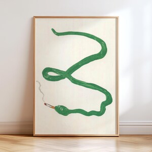 Può includere: Una stampa artistica da parete incorniciata raffigurante un'illustrazione dipinta a mano di un lungo serpente verde sinuoso su sfondo color crema. Il serpente tiene in bocca una piccola sigaretta accesa, con una sottile scia di fumo grigio che sale.