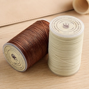 Op de afbeelding: Twee cilindrische klossen naaigaren, een donkerbruine en een crèmekleurige, elk met een rond label aan de bovenkant met de tekst 'Specializing in the manufacture of a variety of handmade sewing thread' en een klein icoontje van een klosje.