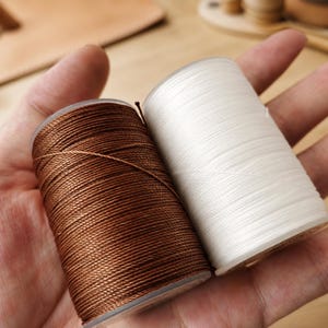 Pode incluir: Dois carretéis cilíndricos de linha, um de cor castanha rica e o outro de cor branca brilhante. Ambos os carretéis estão enrolados firmemente com um cordão sintético liso e durável, com uma textura consistente e um ligeiro brilho na superfície.