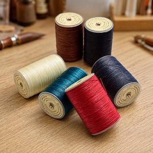 May include: Six spools of waxed thread in various colors including brown, black, cream, teal, red, and dark blue. Each spool features a circular label on the top with printed text and a graphic of a thread spool, arranged in a cluster.