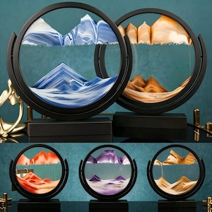 May include: A collection of five circular sand art displays featuring black frames and rectangular bases. Each piece contains colored sand forming layered mountain landscapes in blue, gold, orange, purple, and beige tones within glass.