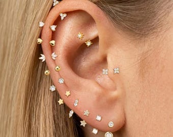 May include: A collection of gold-toned stud earrings arranged along the ear cartilage and lobe. The set includes various shapes such as small stars, hearts, spheres, and clear crystal accents, creating a layered look across the ear structure.