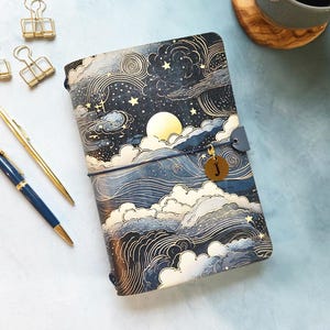 May include: A rectangular journal with a dark blue cover featuring a gold moon, stars, and swirling cloud illustrations. It includes a gold charm with the letter J, two metal pens, and three gold binder clips next to a coffee mug.