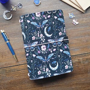 May include: A black journal with a floral and butterfly pattern featuring blue moths, white crescent moons, and pink flowers. The journal is secured with a light pink elastic band and a black tab. A blue ballpoint pen lies next to the journal.