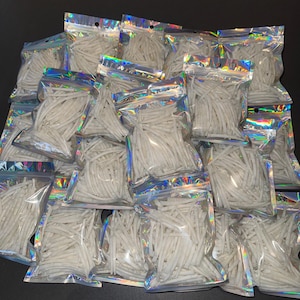 May include: A collection of numerous small, clear plastic bags with holographic edges, each filled with a quantity of thin, white, cylindrical plastic sticks. The bags are arranged in a pile, showing multiple units of the same product.