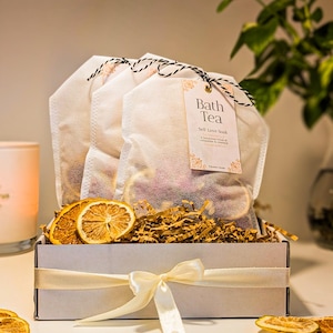 Könnte beinhalten: Eine weiße Geschenkbox mit cremefarbenem Band enthält drei weiße Netzbeutel gefüllt mit getrockneten Pflanzen und Orangenscheiben. Jeder Beutel hat ein Etikett mit der Aufschrift Bath Tea Sell Love Soak. Getrocknete Orangenscheiben liegen im Füllmaterial.