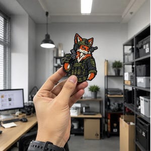 May include: A tactical morale patch featuring a stylised orange and white fox character wearing a green military helmet and tactical vest, holding a black rifle over its shoulder. The design uses bold black outlines and a vibrant colour palette.