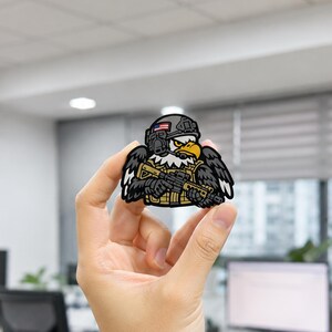 May include: A tactical morale patch featuring a cartoon bald eagle wearing a grey combat helmet with a small American flag decal, a tan tactical vest, and holding a black rifle. The eagle has white feathers on its head and black wings.
