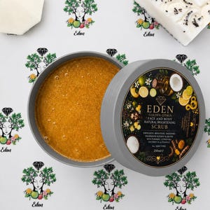 May include: A round grey container of Eden Golden Citrus face and body natural brightening scrub with a golden gel texture. The label features botanical illustrations, turmeric, and citrus. A white soap bar with black seeds is nearby.