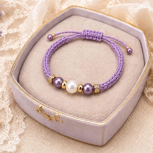 Op de afbeelding: Een armband van gevlochten lavendelkleurig koord met een centrale witte parel geflankeerd door twee paarse parels, goudkleurige metalen tussenstukjes en twee kleinere paarse parels aan de uiteinden van het verstelbare koord.