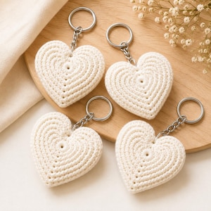 Puede incluir: Cuatro llaveros en forma de corazón de color blanco roto hechos de hilo de ganchillo. Cada corazón presenta un patrón de puntadas circulares concéntricas y está unido a una anilla de metal plateado mediante una cadena corta. Los corazones son suaves y texturizados.