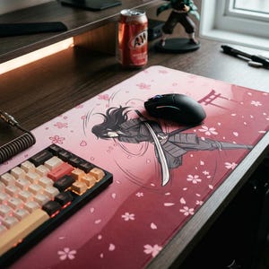 Peut inclure: Un tapis de bureau rectangulaire avec un fond dégradé rose et rouge foncé orné de fleurs de cerisier tombantes. Le motif central représente un guerrier aux longs cheveux en armure sombre maniant un sabre katana.