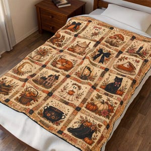 May include: A cream-colored throw blanket featuring a grid pattern of autumn-themed illustrations including pumpkins, a black cat, a ghost, a squirrel, a fox, a house, a lantern, a cauldron, a slice of pie, mushrooms, and floral arrangements.