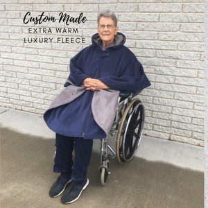 Peut inclure: Cape de fauteuil roulant en polaire bleu marine avec une doublure et une capuche grises. La cape a une fermeture éclair et un bouton au cou. Le texte "Custom Made EXTRA WARM LUXURY FLEECE" est visible.