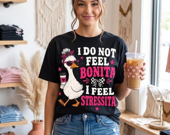 May include: A black short-sleeve t-shirt featuring a graphic of a white duck wearing sunglasses and a pink hair bow, holding a coffee cup. The text reads I DO NOT FEEL BONITA I FEEL STRESSITA in white and pink lettering with small pink stars.