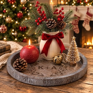 May include: A round wooden tray with a weathered gray finish featuring a laser-engraved wreath and the letters K+A. It holds a white ribbed vase with a red bow, pine branches, red berries, pinecones, a red sphere candle, a bell, and a gold tree.