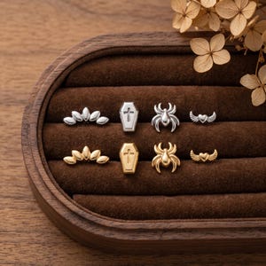May include: A collection of eight small metal stud earrings arranged in two rows. The set includes silver and gold versions of a leaf-like fan, a coffin with a cross, a spider, and a heart with wings, all displayed on a dark brown velvet insert.