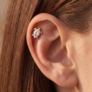 May include: A gold-toned ear cartilage stud shaped like a turtle, featuring a clear, oval-cut gemstone set in the centre of the shell. The metal frame forms the head, four small legs, and a tail, creating a detailed animal-inspired jewellery piece.