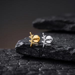Könnte beinhalten: Zwei Ohrstecker mit flacher Rückseite und einem zweigartigen Design mit kleinen Blättern. Ein Ohrstecker ist in poliertem Goldton gehalten, während der andere in poliertem Silberton ausgeführt ist. Jeder Stecker hat eine runde, flache Basis.