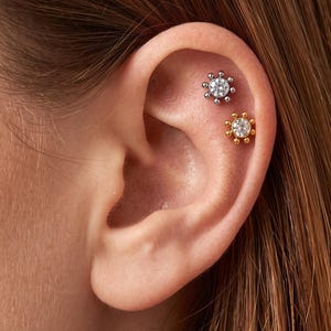 Può includere: Due orecchini da cartilagine a forma di sole sono indossati nella parte superiore dell'orecchio. L'orecchino superiore è color argento con una pietra chiara centrale circondata da piccole sfere metalliche. Quello inferiore è dorato e identico.