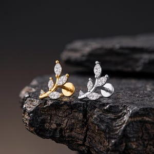 Könnte beinhalten: Zwei Ohrstecker in Form kleiner Zweige, einer in Gold- und einer in Silberoptik. Jeder Ohrstecker ist mit fünf klaren, marquise-geschliffenen Steinen besetzt, die wie Blätter angeordnet sind, und verfügt über einen flachen, runden Verschluss.