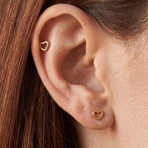 May include: Two gold-toned heart-shaped stud earrings. One earring is positioned in the upper cartilage area of the ear, while the second earring is placed in the lower earlobe. Both pieces feature a simple, open-frame heart design.