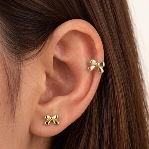 May include: A pair of gold-toned earrings featuring a bow design. One earring is a stud positioned on the earlobe, while the other is a cuff style earring attached to the upper cartilage of the ear. Both pieces have a polished, smooth finish.