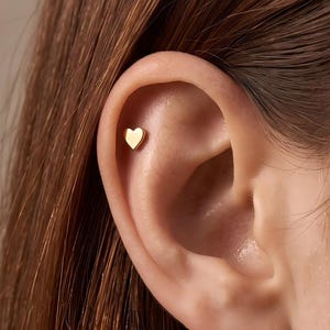 Könnte beinhalten: Ein kleiner, polierter, herzförmiger Ohrstecker in Goldoptik befindet sich im oberen Knorpelbereich des Ohrs. Der Ohrstecker hat eine glatte, reflektierende Oberfläche und ein schlichtes, klares Design, das flach auf der Haut aufliegt.