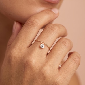 May include: A gold chain ring featuring a single round, clear, faceted gemstone charm suspended from the centre. The delicate metal chain links form the band of the ring, while the small, brilliant-cut stone hangs freely from a small jump ring.