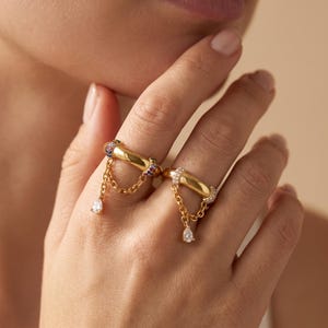 Op de afbeelding: Twee goudkleurige ringen gedragen aan aangrenzende vingers, elk met een dikke band, een bungelend kettinkje en een druppelvormige steen. De ene ring heeft veelkleurige stenen op de band, de andere ring is bezet met heldere stenen.