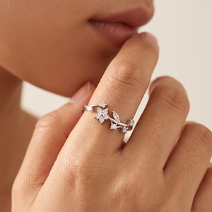 Puede incluir: Un anillo de tono plateado con una delicada banda en forma de enredadera adornada con pequeñas piedras transparentes dispuestas en un patrón floral, que incluye una flor central de cinco pétalos, detalles en forma de marquesa y piedras redondas.