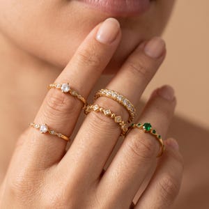May include: A collection of five gold rings worn on fingers. Two rings feature a single clear stone with a delicate band, one ring has a row of clear stones, one ring has a geometric square pattern with small stones, and one ring has a green stone.