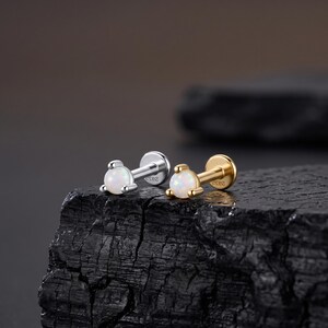 May include: Two flat-back labret studs featuring round, iridescent white opal cabochons held by three prongs. One stud is silver-toned and the other is gold-toned, both displaying the S925 hallmark on the post, designed for body piercings.