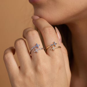 Puede incluir: Dos anillos de estilo abierto con motivos de cruz. Un anillo es de tono plateado con piedras azules y transparentes, mientras que el otro es de tono dorado con piedras azules y transparentes. Cada anillo tiene dos extremos en forma de cruz.