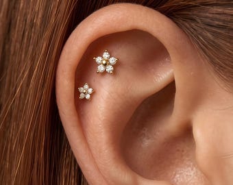 Puede incluir: Dos pendientes dorados con forma de flor que presentan piedras transparentes de corte redondo dispuestas como pétalos alrededor de una cuenta central dorada. Los pendientes están situados en el cartílago superior de la oreja.