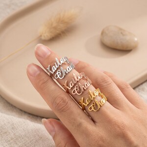 Pode incluir: Três anéis empilhados em tons de prata, ouro rosa e ouro, cada um com os nomes personalizados Kailey e Chari em uma fonte cursiva. Os anéis são usados em um dedo, exibindo o acabamento metálico e o design das letras personalizadas.